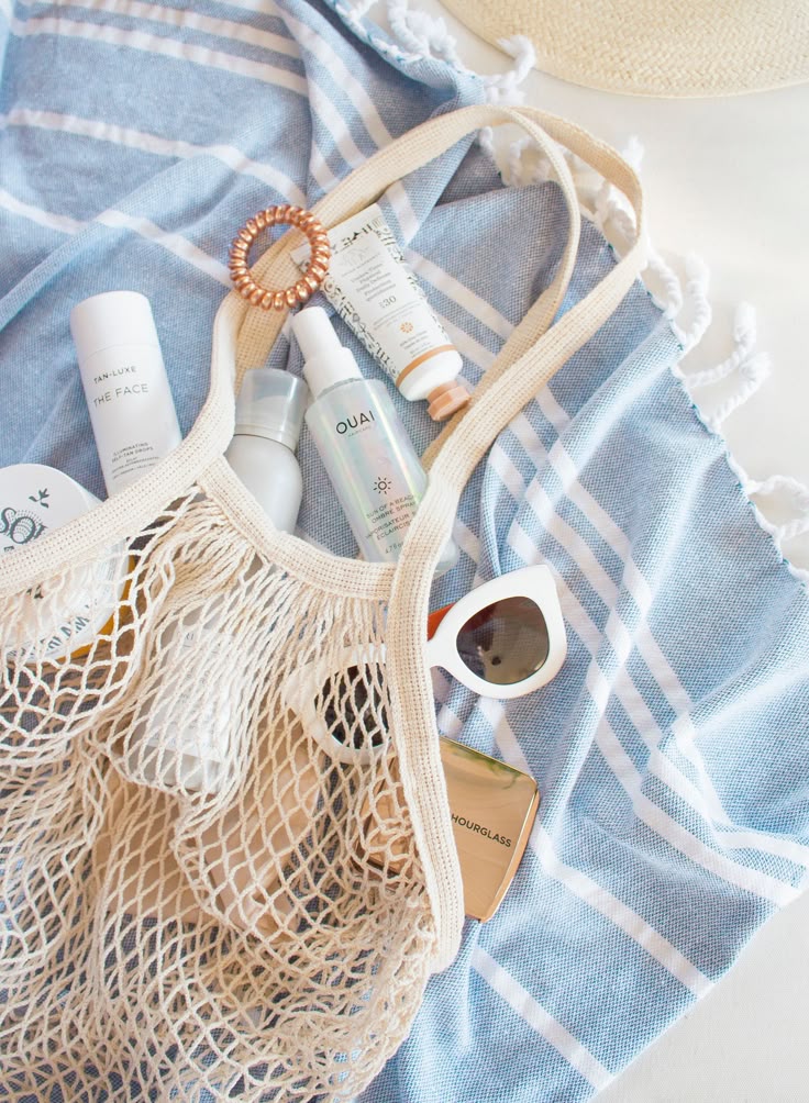 Beach essentials in a net bag: skincare products, sunglasses on a blue striped towel, ready for summer relaxation.