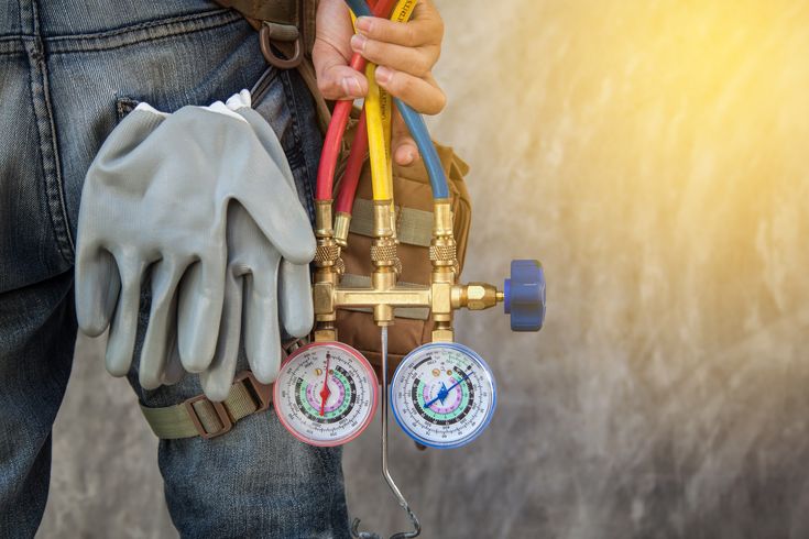 Technician holding HVAC manifold gauges and gloves, sunlight in background.