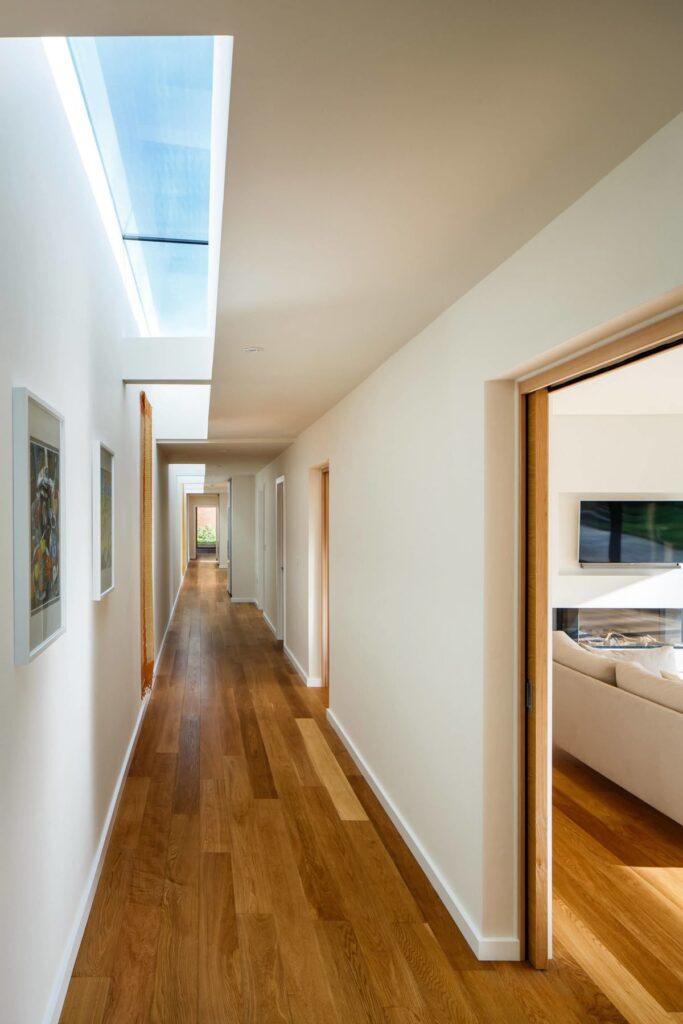 Modern hallway with wooden floors and natural light from skylights, leading to rooms with framed art on walls.