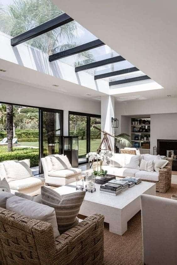 Modern living room with skylight, wicker chairs, white sofas, and garden view through large windows.
