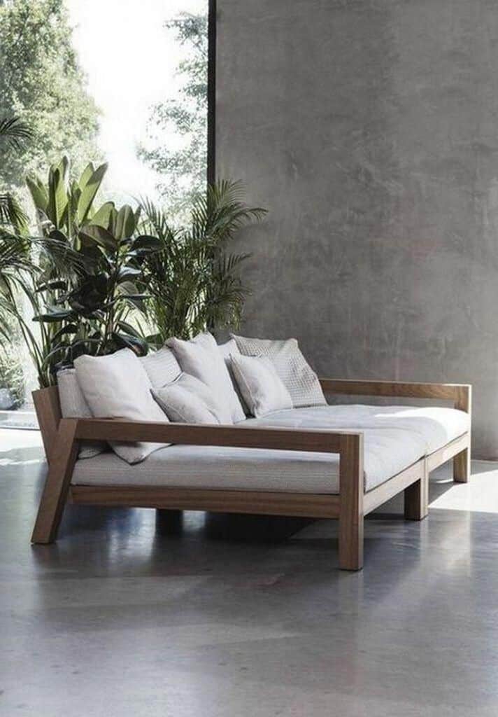 Modern wooden daybed with white cushions, surrounded by green plants in a bright, minimalist room.