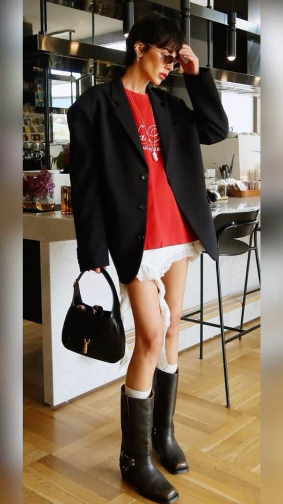 Fashionable woman in oversized blazer and boots, holding a black handbag, posing in a modern kitchen space.