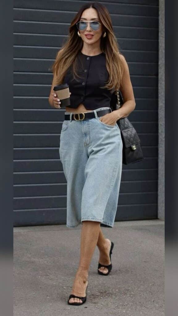 Woman in chic summer outfit with denim shorts, holding coffee, wearing sunglasses, walking outdoors.