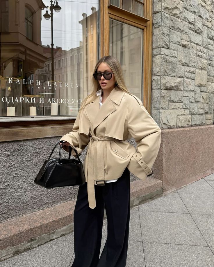Stylish woman in trench coat and sunglasses holding a black handbag, standing near a store window display.