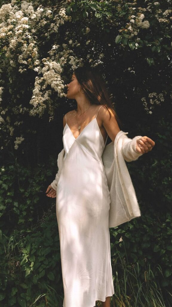 Woman in white dress stands near blooming bushes, basking in sunlight.