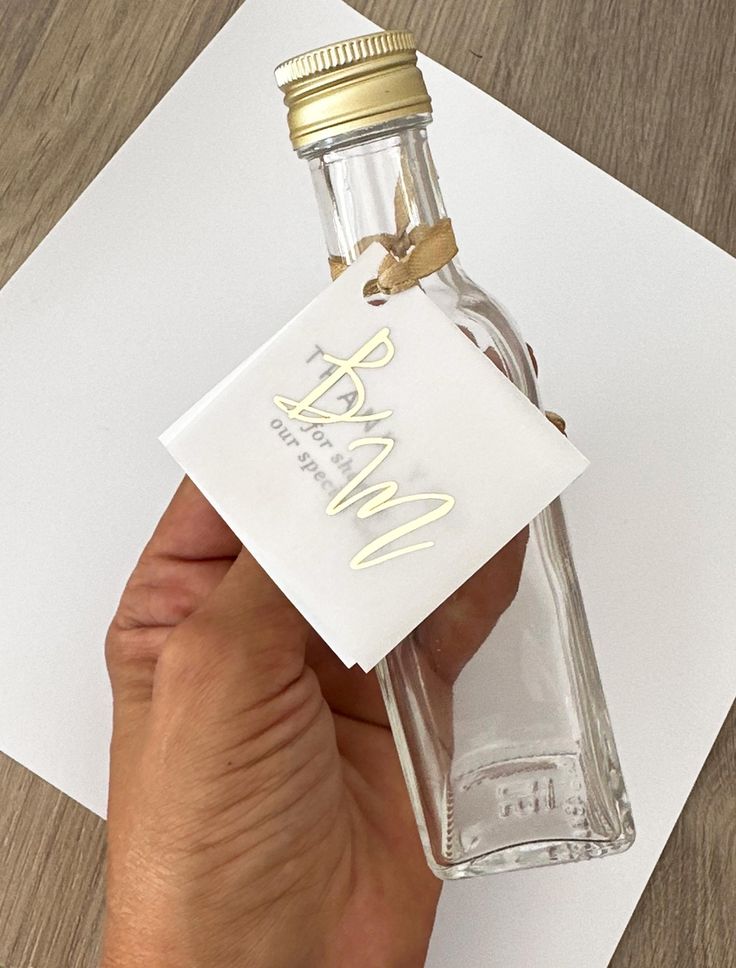 Clear glass bottle with gold cap and personalized tag, held by hand, on a wooden surface.