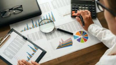 Person analyzing financial charts with magnifying glass, calculator, and colorful graphs on a desk.