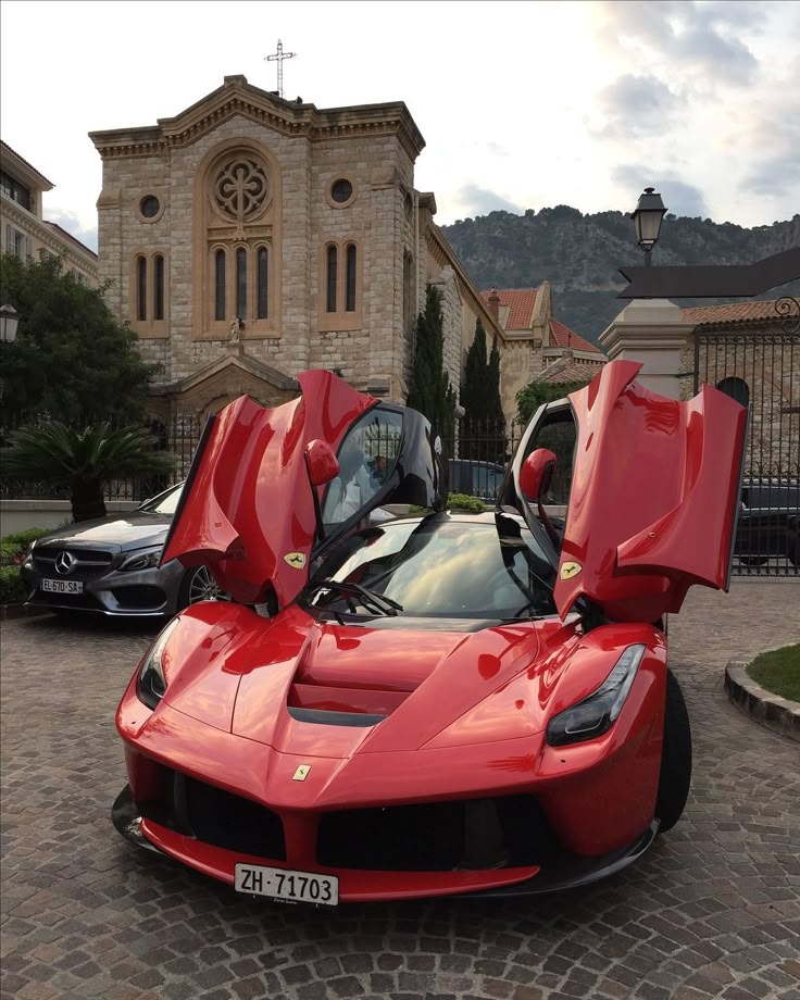 Red sports car with scissor doors open, parked on a cobblestone street in front of an elegant stone building.