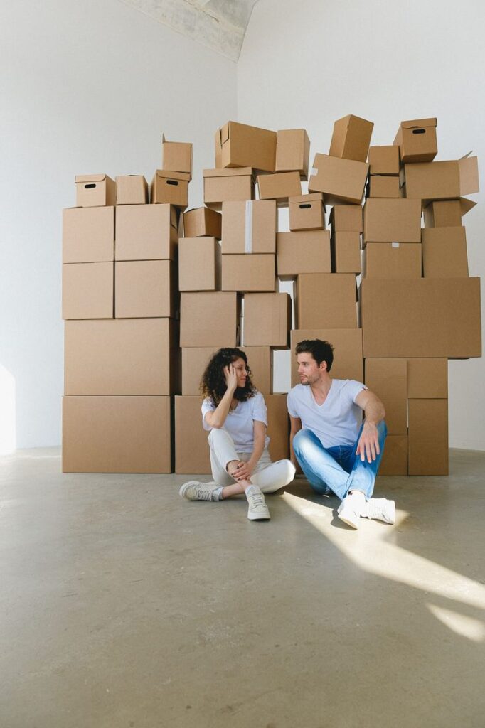 Couple sitting on floor surrounded by packed moving boxes in a bright room.