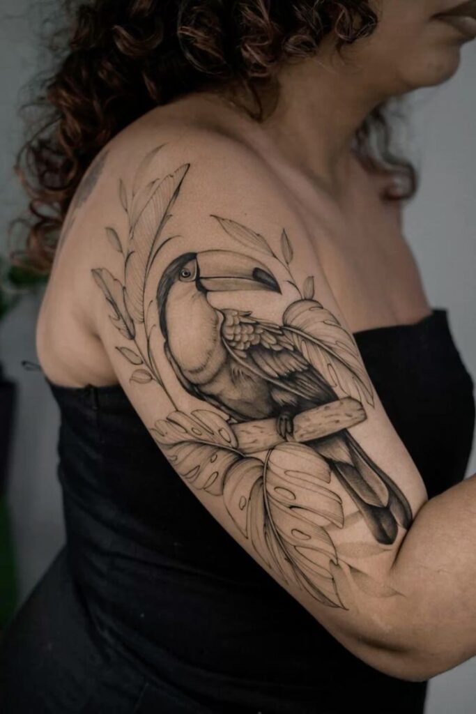 Toucan tattoo with floral design on a woman's upper arm, showcasing intricate black and gray details.