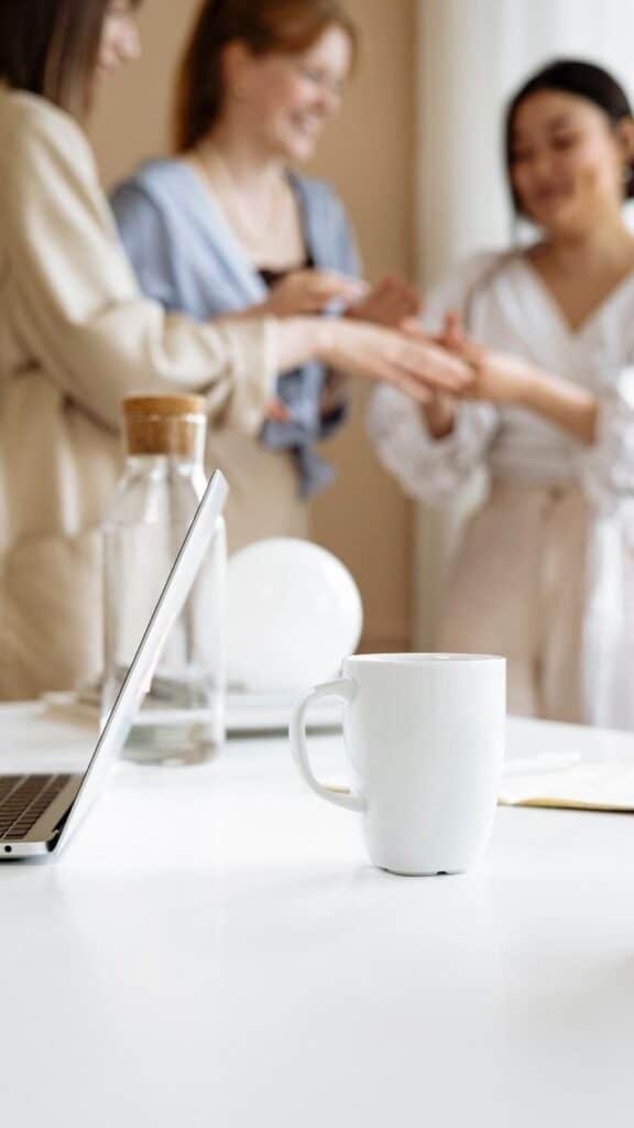 Laptop and coffee mug on office desk with people chatting in the background.