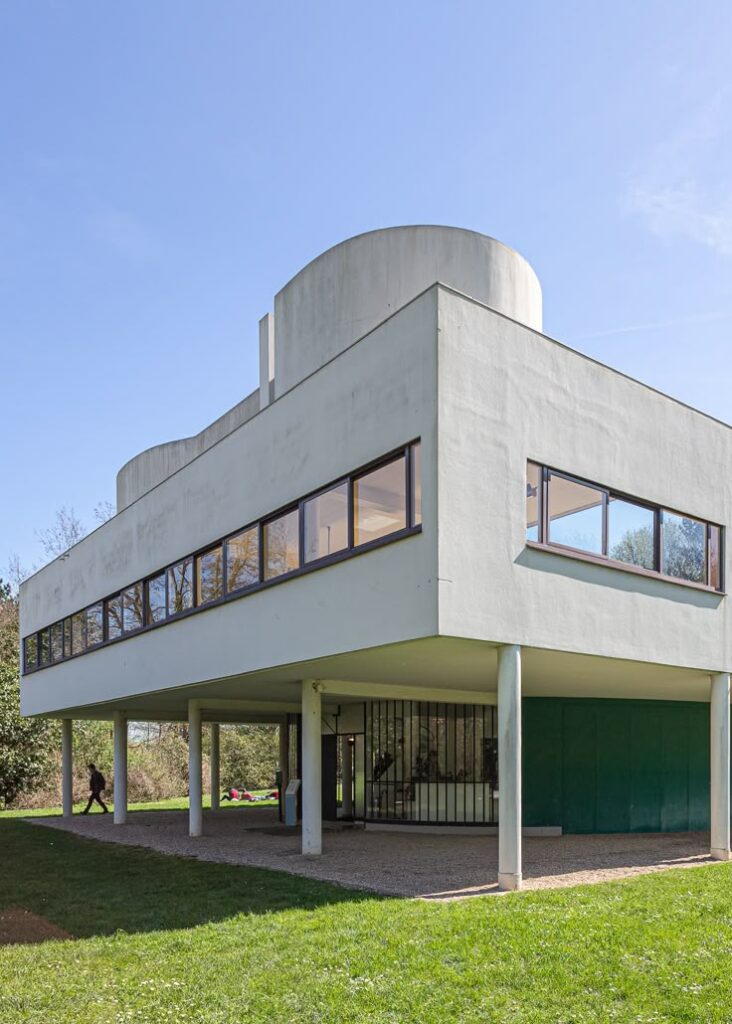 Modernist white building on stilts with large windows, set in a green landscape under a clear blue sky.