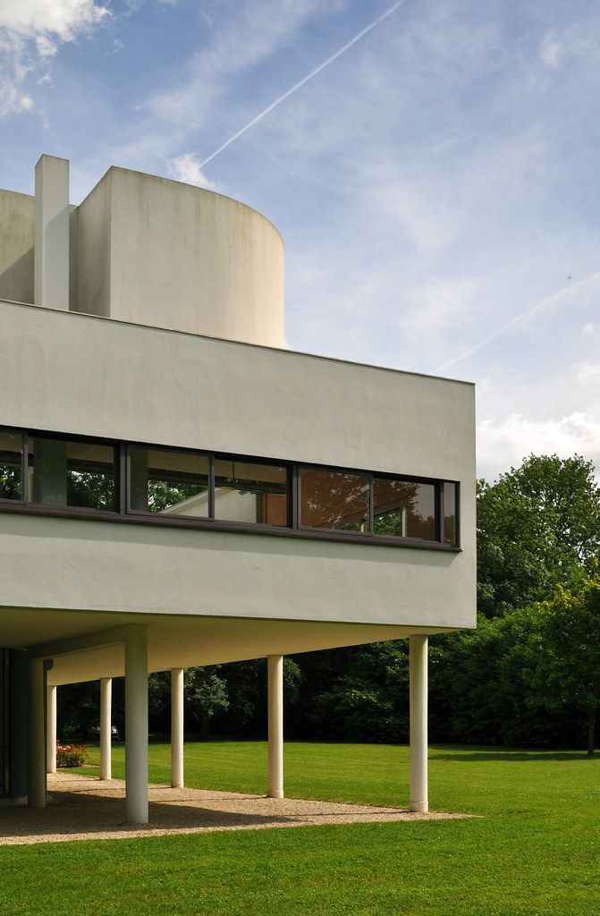 Modernist building with white facade, large windows, and stilts, set in a green landscape under a blue sky.