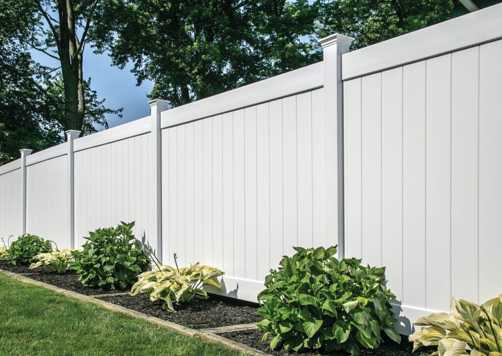 White vinyl fence with lush green plants and trees in a backyard setting.