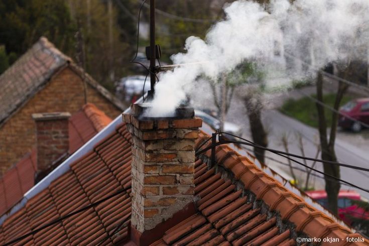 Smoke billowing from a red brick chimney on a tiled roof, with blurred street and houses in the background.