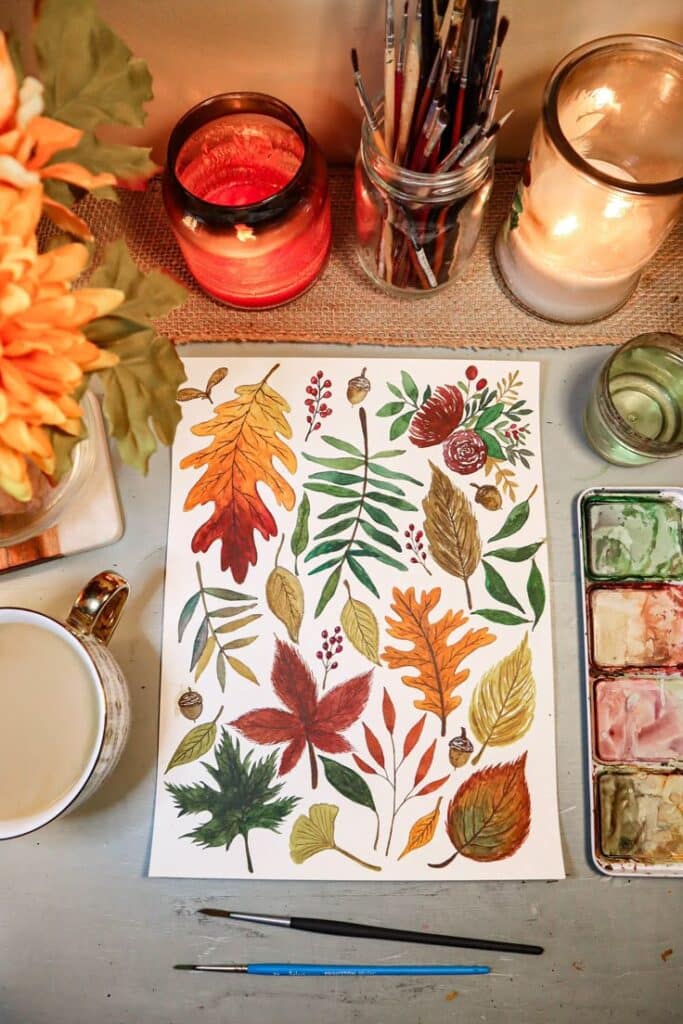 Watercolor leaf painting with autumn colors, surrounded by candles, paintbrushes, and a mug on a table.
