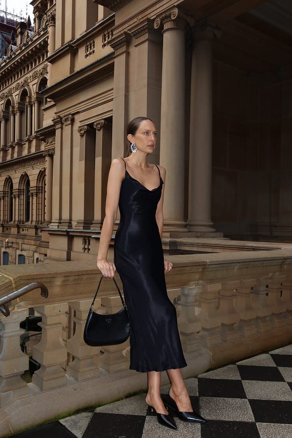 Woman in elegant black dress with handbag poses outside historic building.