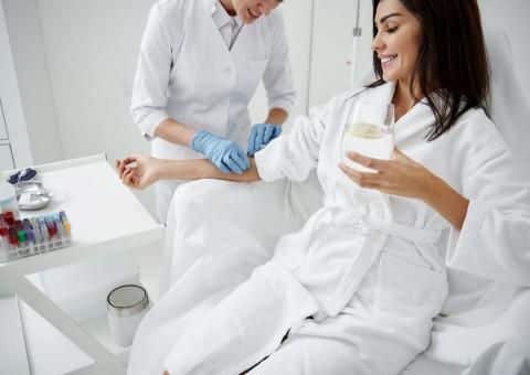 Woman receiving IV therapy in clinic while holding a glass, attended by healthcare professional in white coat and gloves.