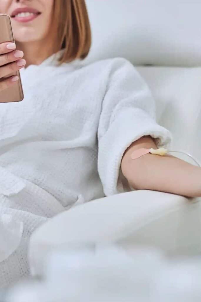 Person relaxing with IV drip, holding a smartphone, in a health spa setting.