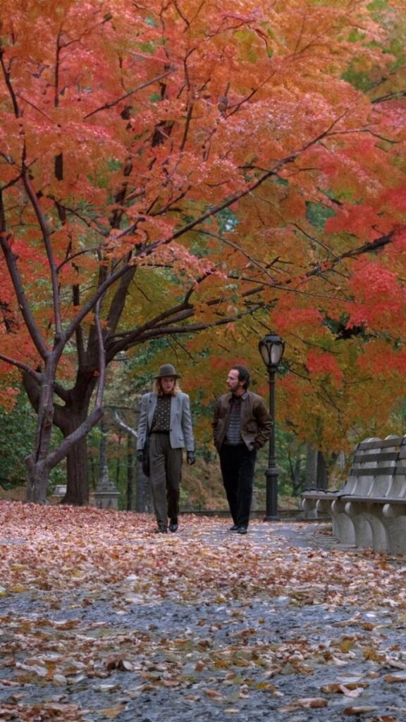 Couple walking under vibrant autumn leaves in a scenic park, surrounded by fallen foliage and benches.