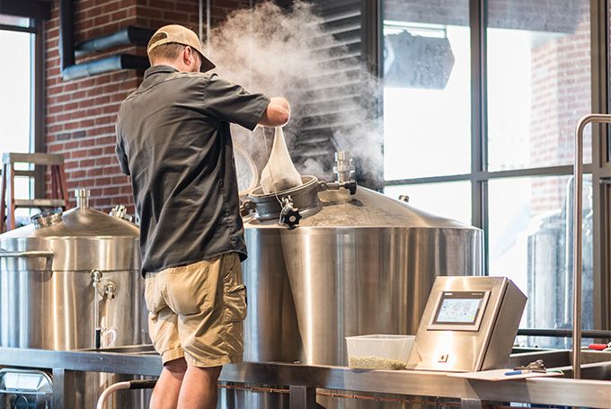Brewer adding ingredients to steaming tank in a brewery, surrounded by equipment and controls.
