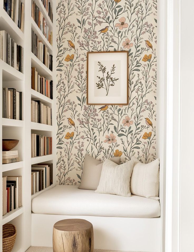 Cozy reading nook with floral wallpaper, white shelves filled with books, and a soft bench with neutral cushions.