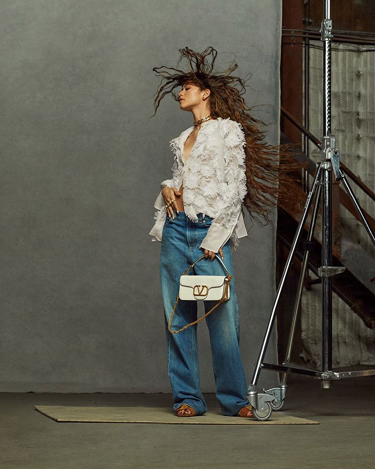 Woman in a stylish outfit with a white blouse and blue jeans, holding a designer handbag in a studio setting.