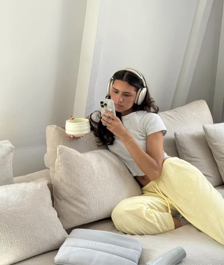 Woman lounging on a sofa with headphones, holding a phone and cup, wearing casual clothes, enjoying a relaxed moment.