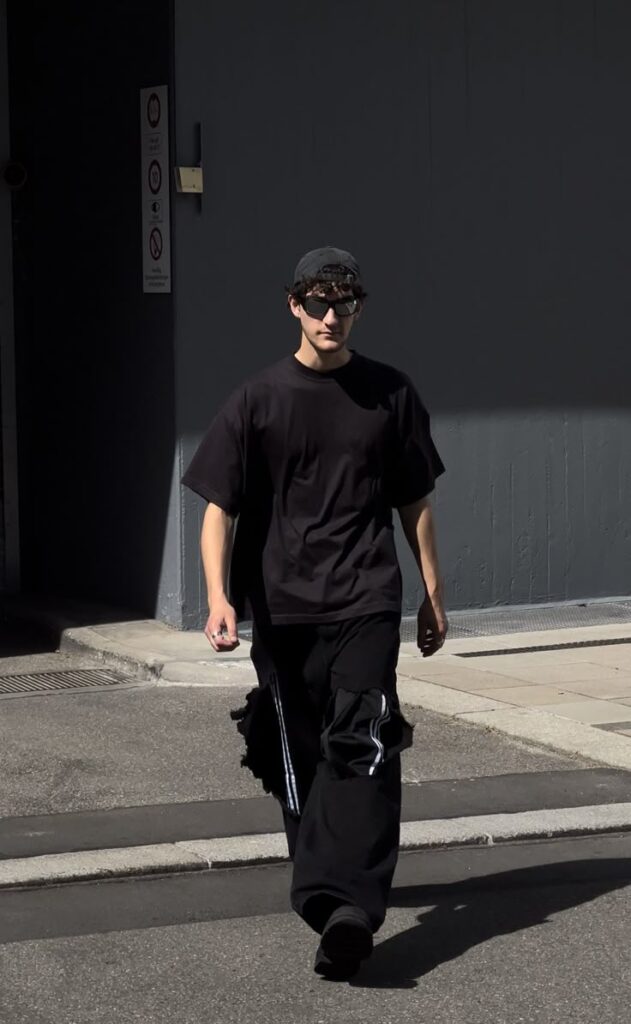 Person in black outfit and sunglasses walking on street in urban setting, casting a shadow on sunny day.