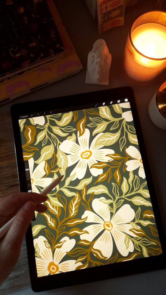 Tablet displaying floral digital art with stylus, surrounded by candles and books, on a cozy desk setup.