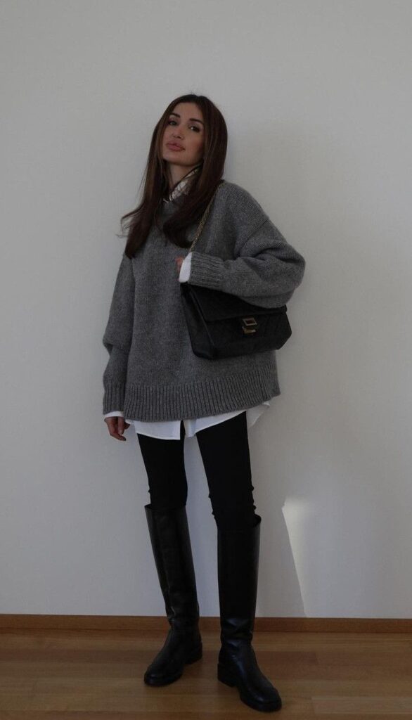 Woman in stylish outfit with gray sweater, white shirt, black leggings, tall boots, and a quilted handbag.