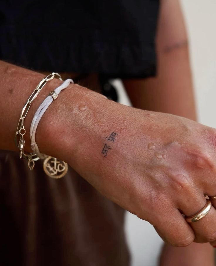 Close-up of a hand with a small tattoo and gold jewelry, featuring water droplets for a fresh look.