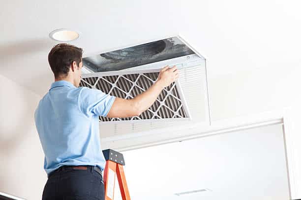 Person replacing ceiling air filter on ladder, performing maintenance in a home interior.