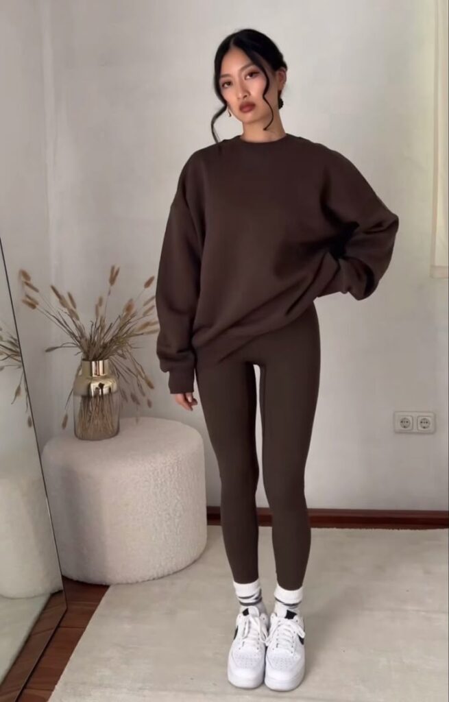 Person in brown outfit with white sneakers stands in a minimal room, near a mirror and a decorative plant.