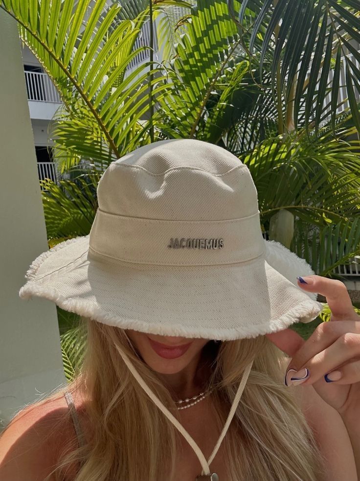 Woman in a stylish beige bucket hat with greenery in the background, shielding face from the sun.