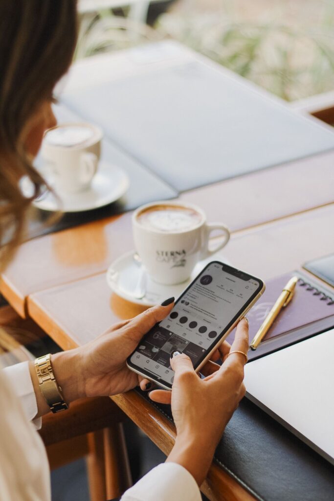 Woman browsing phone at café table with coffee and notebook nearby.