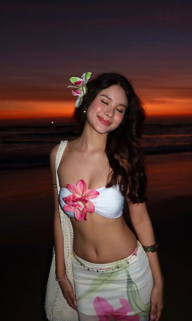 Woman smiling at sunset beach, wearing floral attire, with flower in hair. Relaxation and tropical vibes.