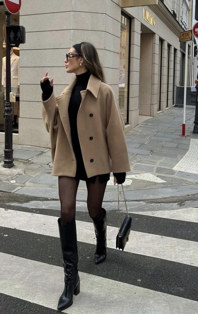 Stylish woman crossing street wearing a beige coat and black knee-high boots, holding a chic handbag.