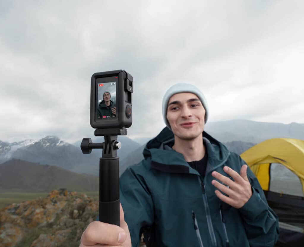 Man filming vlog with camera on selfie stick in scenic mountain landscape near yellow tent.