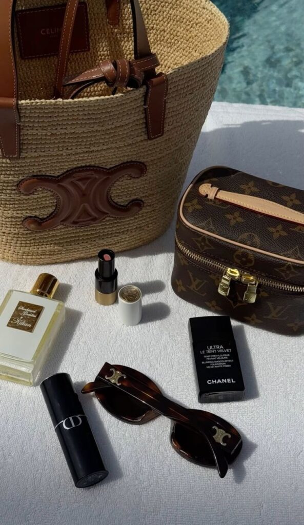 Beach essentials: straw tote, designer cosmetic bag, sunglasses, perfume, lipstick, foundation on towel by poolside.