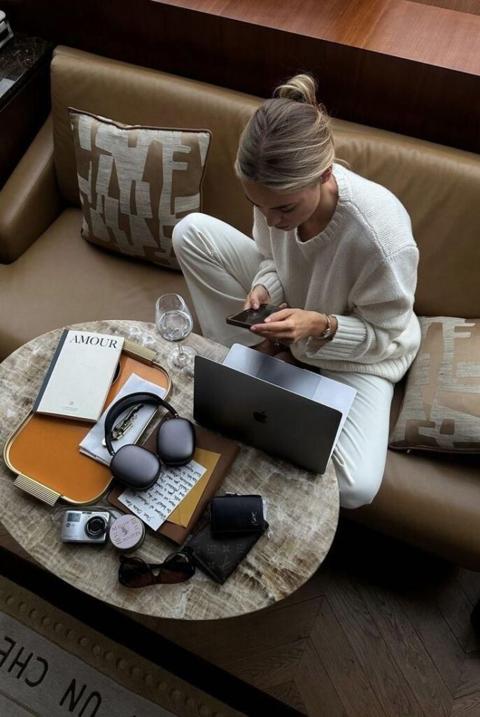b9072cb44b3ab9f5fbdb57a2a0fcbb07 Woman in cozy setting using phone and laptop, surrounded by magazines, headphones, camera, and sunglasses on a table. | Sky Rye Design Woman in cozy setting using phone and laptop, surrounded by magazines, headphones, camera, and sunglasses on a table.