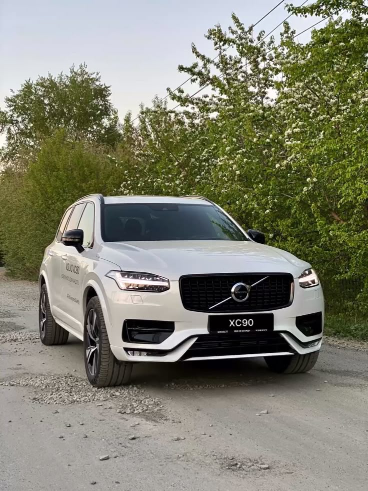 White Volvo XC90 parked on a gravel road, surrounded by green trees in daylight.