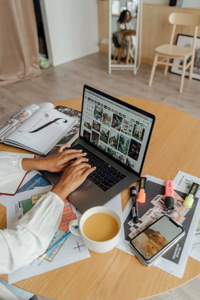 Person working on a laptop surrounded by magazines, highlighters, and coffee, featuring interior design inspiration.