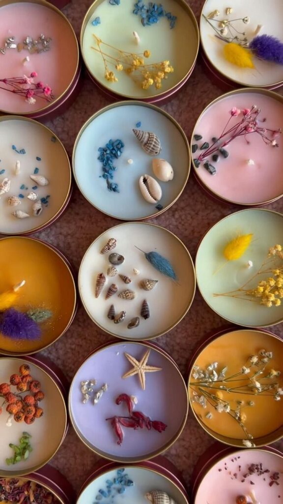 Colorful candles with dried flowers, seashells, and decorative elements in round tins, arranged in rows.