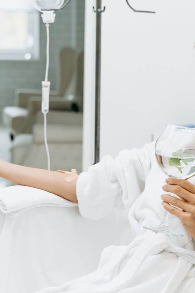 bbb9880f501ee41339792e43eaef134e Person in a robe receiving IV therapy while holding a glass of infused water in a spa setting. | Sky Rye Design Person in a robe receiving IV therapy while holding a glass of infused water in a spa setting.