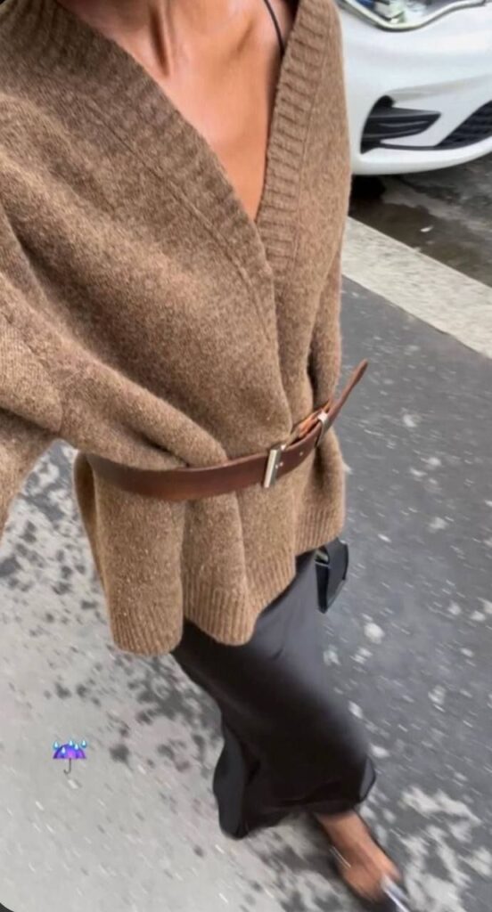 Person walking in oversized brown sweater belted at the waist, paired with a black skirt and pointed shoes.