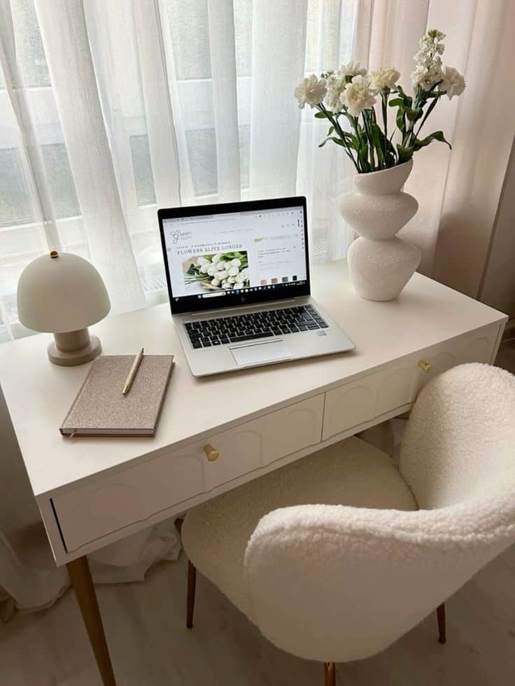 Cozy home office setup with laptop, notebook, table lamp, and flowers on a white desk by a window with sheer curtains.