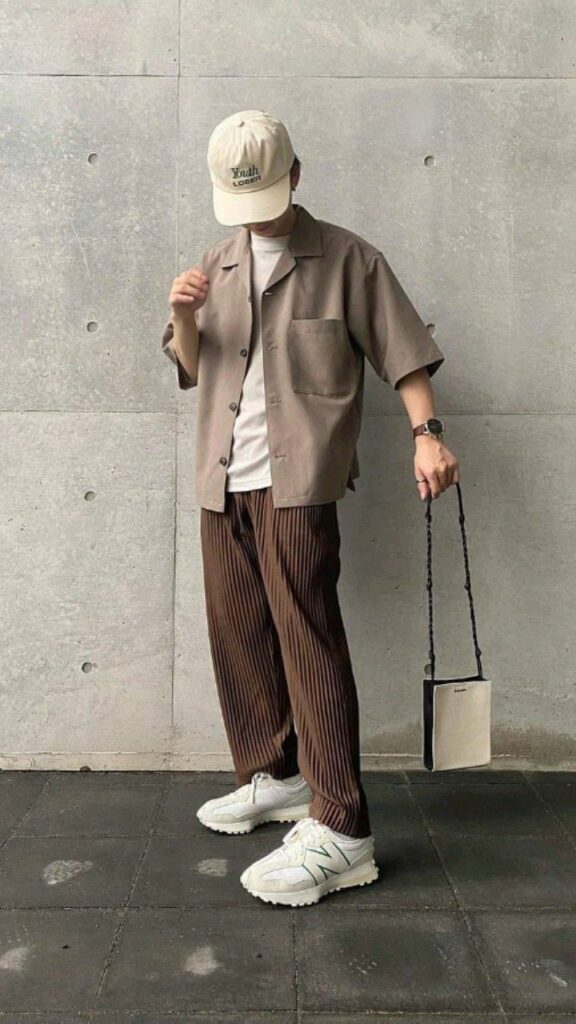 Person in casual streetwear with cap and handbag, standing against a concrete wall.