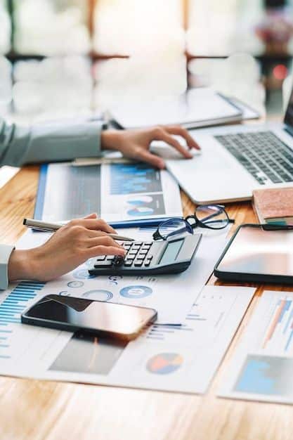 Person using a calculator at a desk with charts, laptop, and phone, analyzing financial data for business insights.