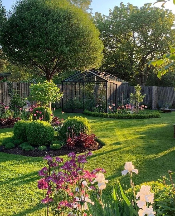 Lush garden with greenhouse, colorful flowers, and vibrant greenery under a clear sky. Perfect spring garden scene.